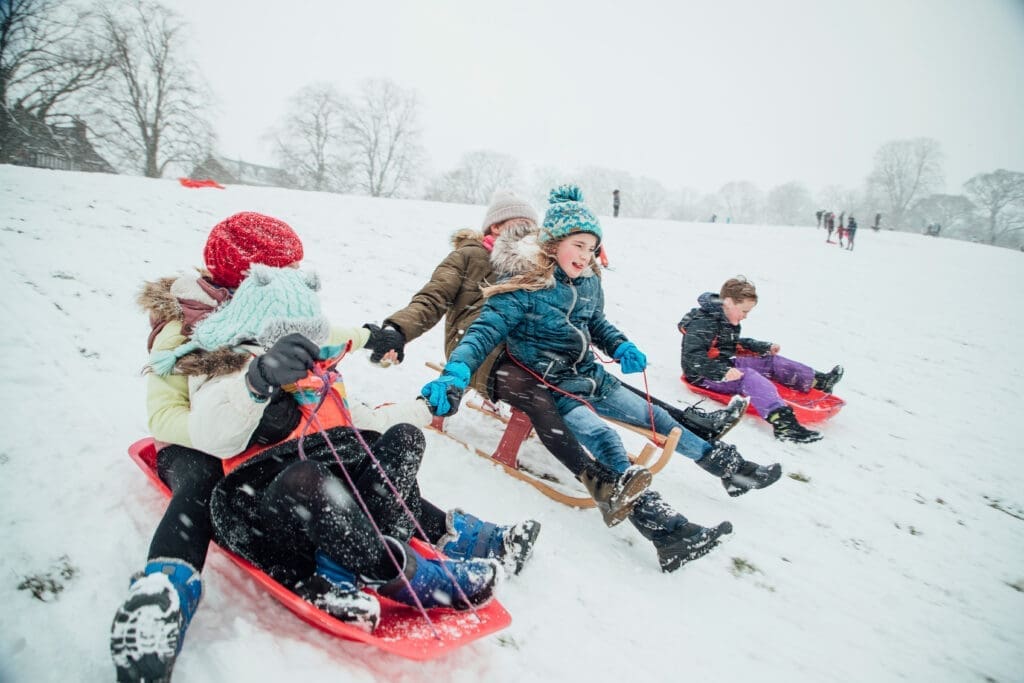Children are racing each other down a hill on sleds in the snow. The girls are holding hands.